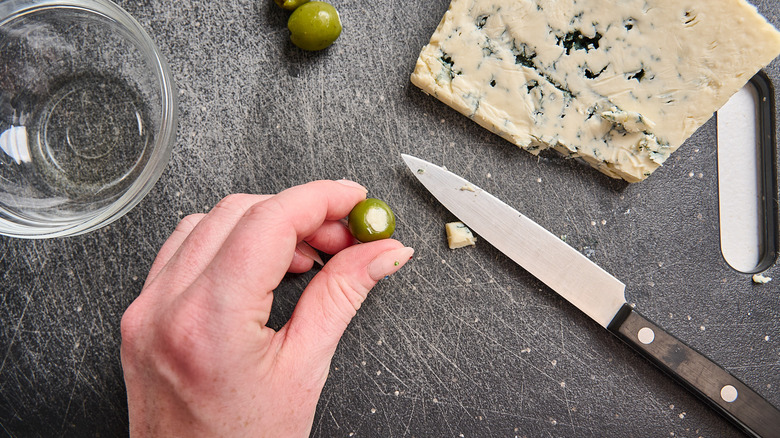 hand holding cheese stuffed olive next to knife and block of blue cheese