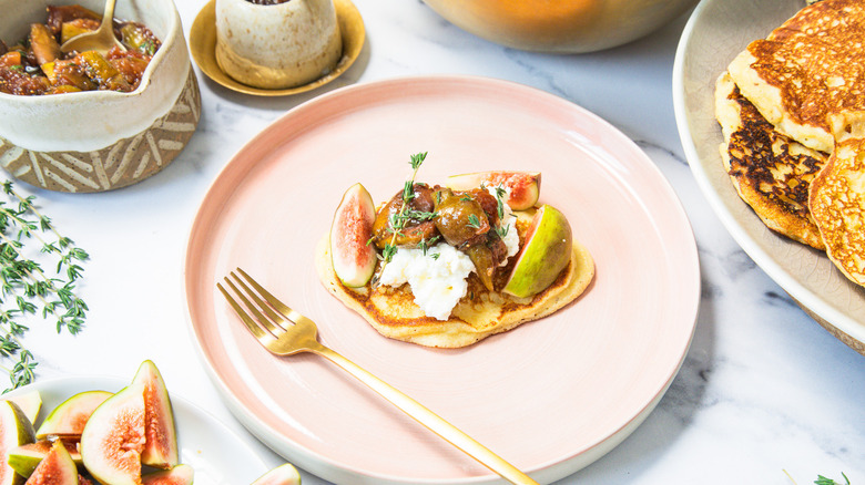 A table scene of one goat cheese pancake on a plate with all the toppings and pancakes around.