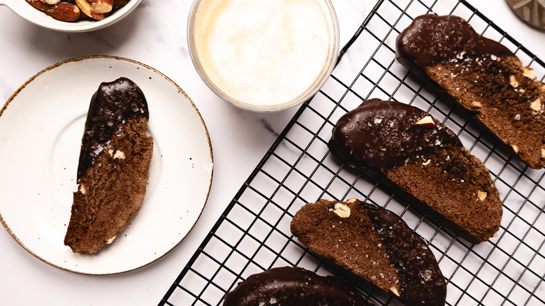 Four chocolate biscotti on a rack with ganache in background, and one on a plate.