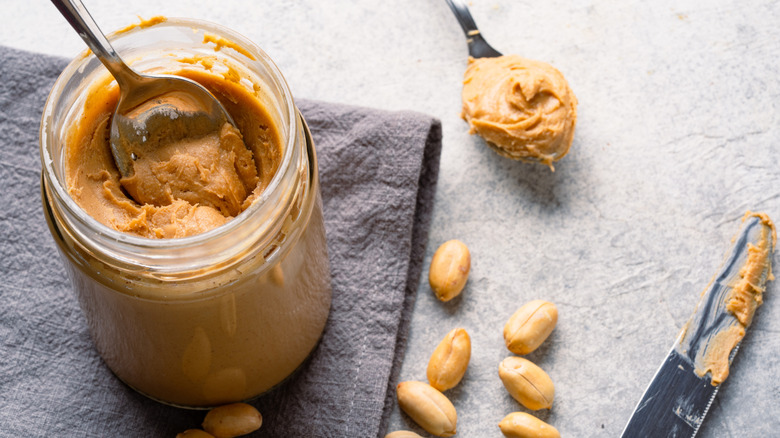 Jar of peanut butter with peanuts and utensils on a counter
