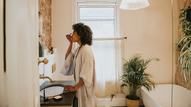 A beautiful black woman stands in a stylish bathroom in a long, silky robe and carefully applies her make-up.