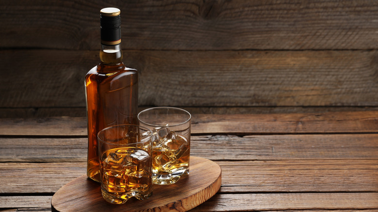 Glass bottle of bourbon next to two rocks glasses full of ice and bourbon against wooden board backdrop