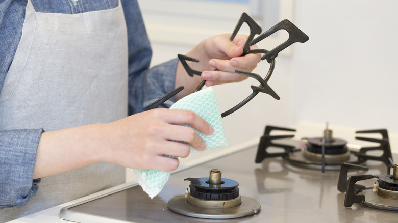 A person in a gray apron cleaning a black gas stove element with a cloth.