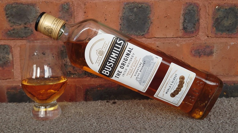 A bottle of Bushmills Original leaning on the rim of a filled whiskey glass
