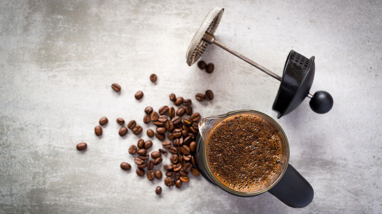 Top view of an open French press filled with coffee, alongside the plunger and whole coffee beans.