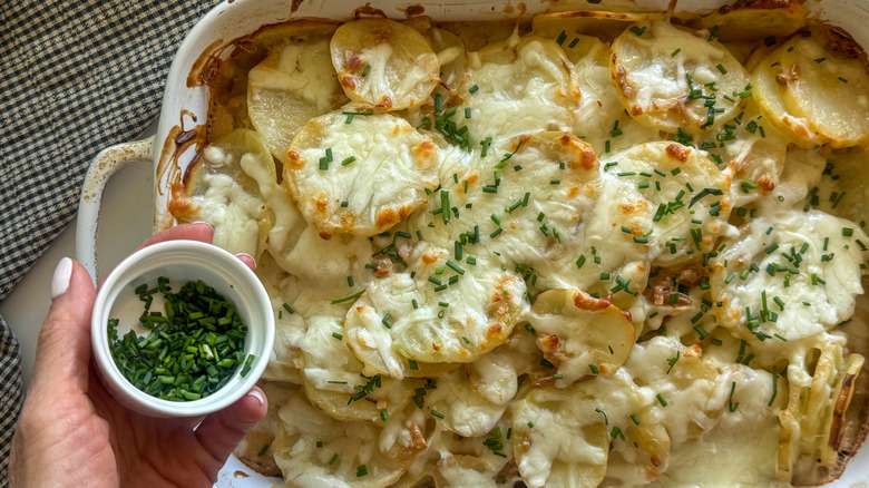 hand adding chives to cheesy potatoes in casserole dish