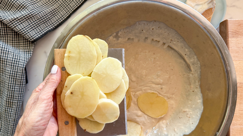 hand adding sliced potatoes to cream mixture in bowl