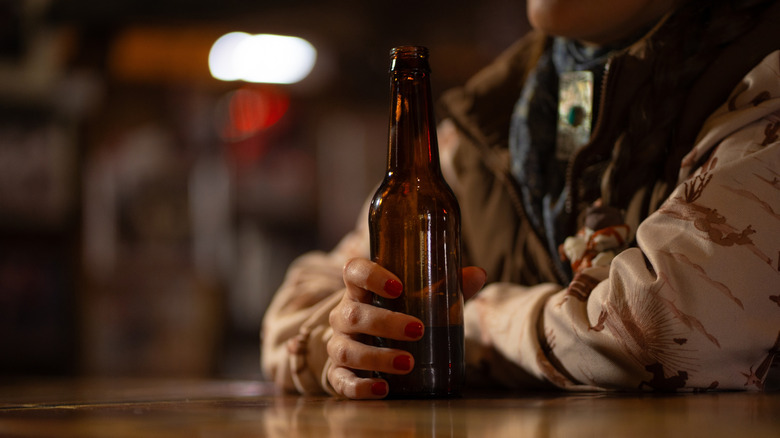 Person in Western garb sitting a bar with a brown glass bottle in one hand
