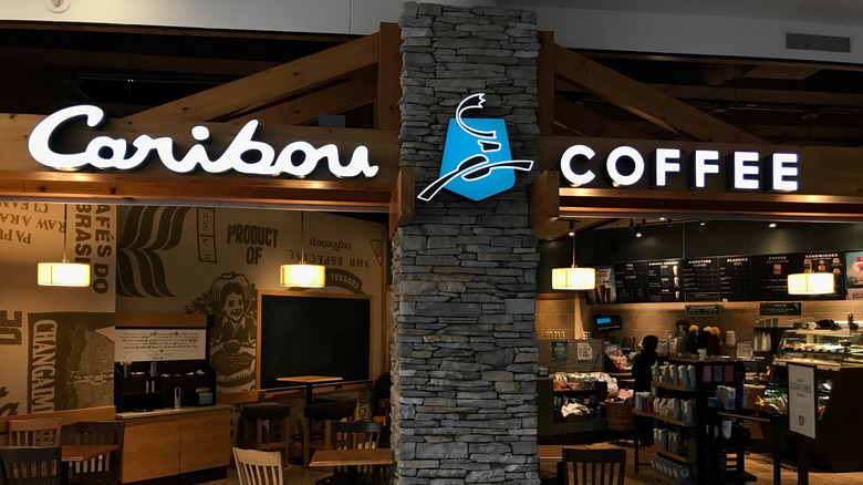 The Caribou Coffee storefront, featuring the brand's name, as well as tables, chairs, and coffee cups for purchase, are pictured inside a mall.