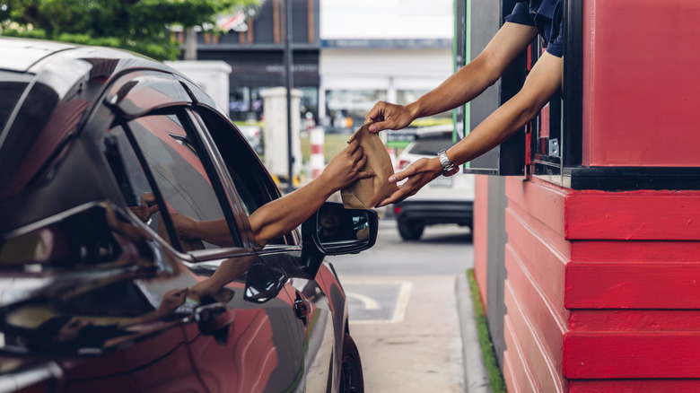 A passenger in a black car receives a brown take-out bag from an employee's outstretched hands in a drive-through.