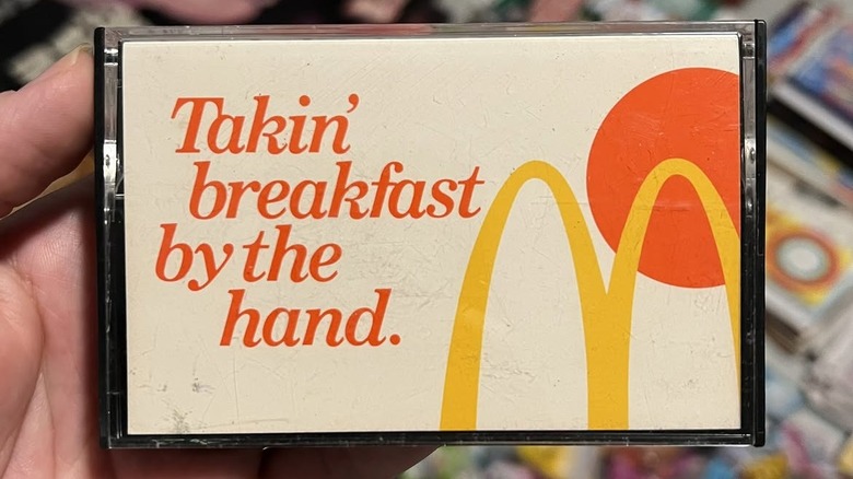 a hand holding a cassette titled "Takin' Breakfast By The Hand," featuring McDonald's golden arches