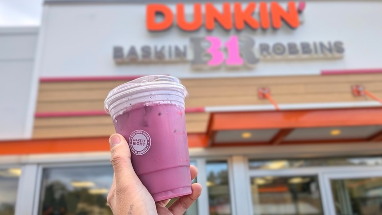 A hand holding a purple Dunkin' refresher in front of a Dunkin'-Baskin Robbins storefront