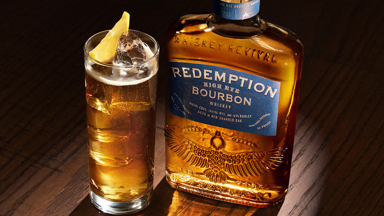 A high-to-low picture of a bottle of Redemption High Rye Bourbon next to a cocktail with light shining though them