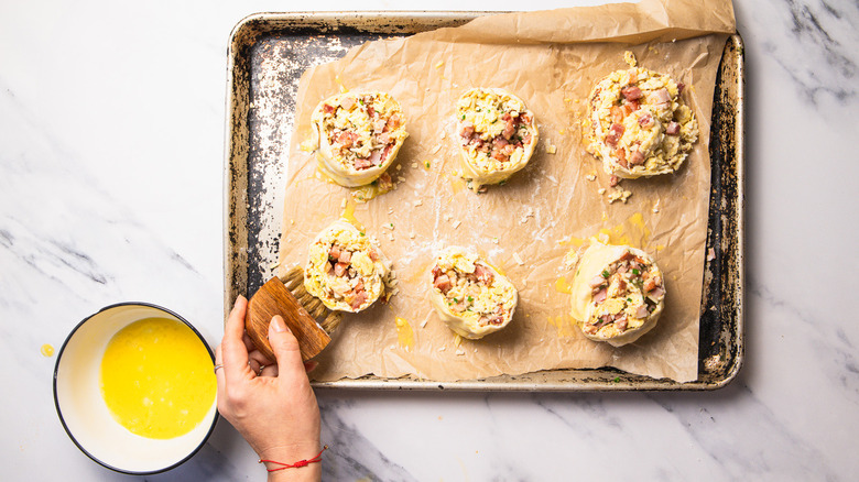 brushing stuffed eggs Benedict rolls with egg wash on baking sheet